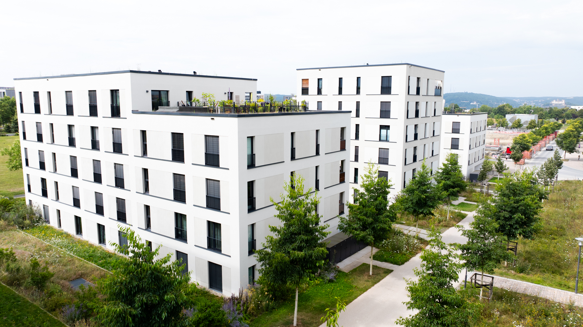 Wohnhäuser mit einer Drohne fotografiert. Die Häuser sind weiß und wirken sehr modern. Neben großen Fenstern sieht man eine Dachterrasse und viel Wiese und Bäume außen herum bepflanzt.